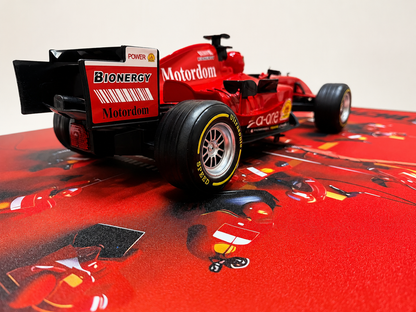 A red and black scale model of a Formula 1 car, resembling a Scuderia Ferrari vehicle with visible Bionergy and Motodom sponsor logos, displayed on a red printed surface.